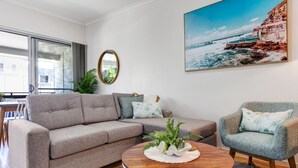 A modern living room with a grey sofa, a round wooden coffee table with a decorative plant, and a framed coastal landscape painting. - MARJ12- Stylish and Spacious Apartment  (Mooloolaba)