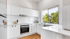 A modern kitchen with white cabinetry, a stainless steel oven, and a window with a view of greenery and buildings. - MARJ12- Stylish and Spacious Apartment  (Mooloolaba)