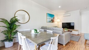 A modern living room with a grey sofa, a dining table with white chairs, a large mirror, and a potted plant. - MARJ12- Stylish and Spacious Apartment  (Mooloolaba)