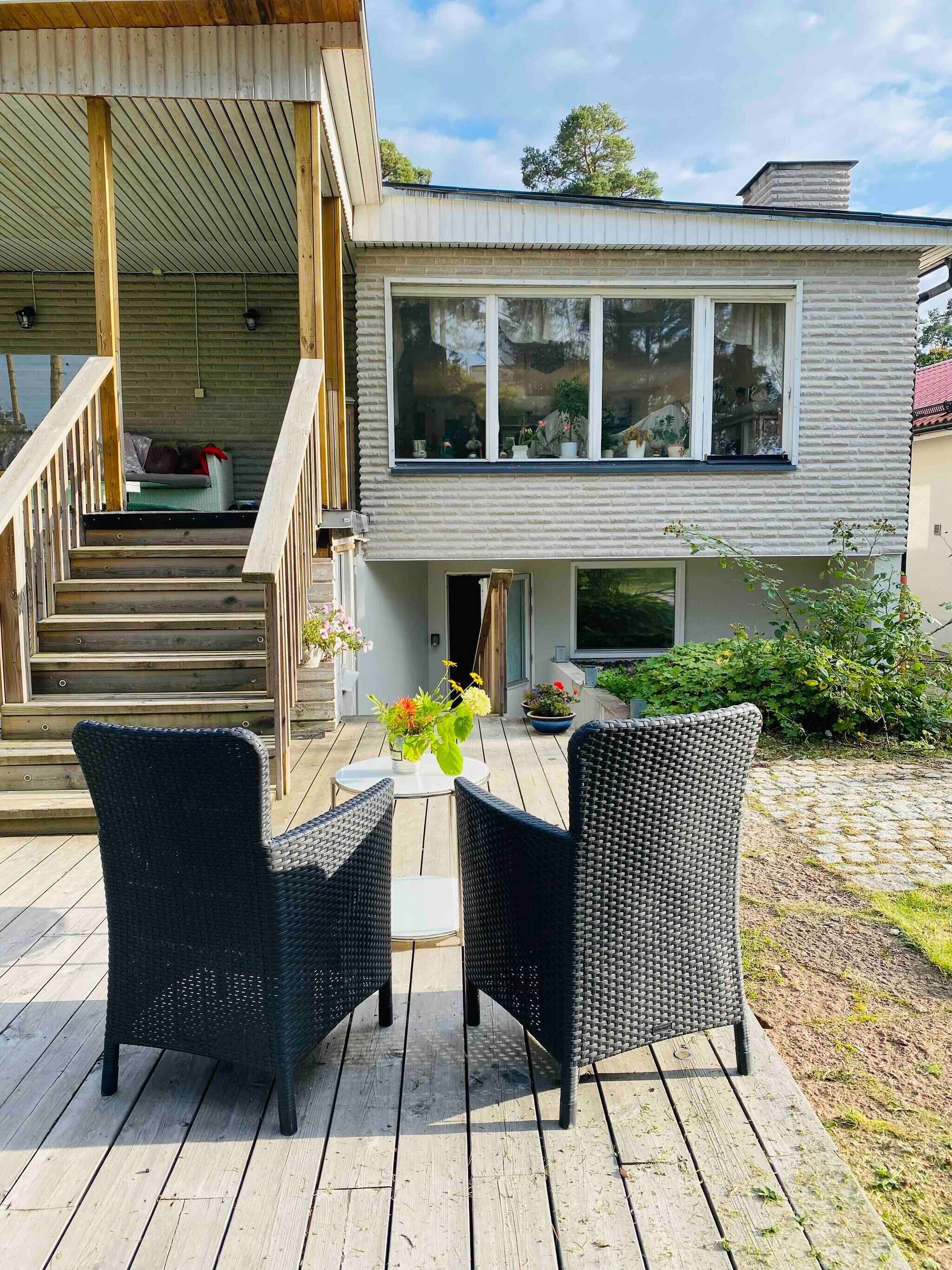 A modern house with a wooden deck, two wicker chairs, a small table with a vase of flowers, and a staircase leading to a covered porch.