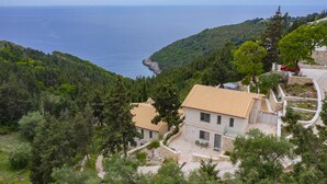A stone house with a tiled roof, surrounded by lush greenery and a coastal view. - Prenari Villa in Paxi (Paxos)