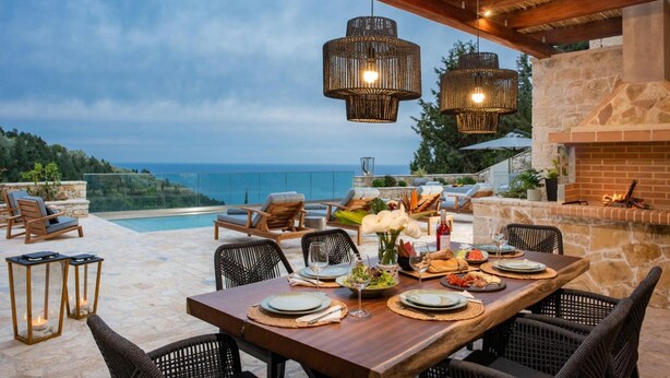 An outdoor dining area with a view of the sea, wicker chairs, a wooden table set for a meal, and hanging wicker light fixtures. - Prenari Villa in Paxi (Paxos)
