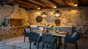 An outdoor dining area with stone walls, a brick fireplace, wicker chairs, and hanging light fixtures. - Prenari Villa in Paxi (Paxos)