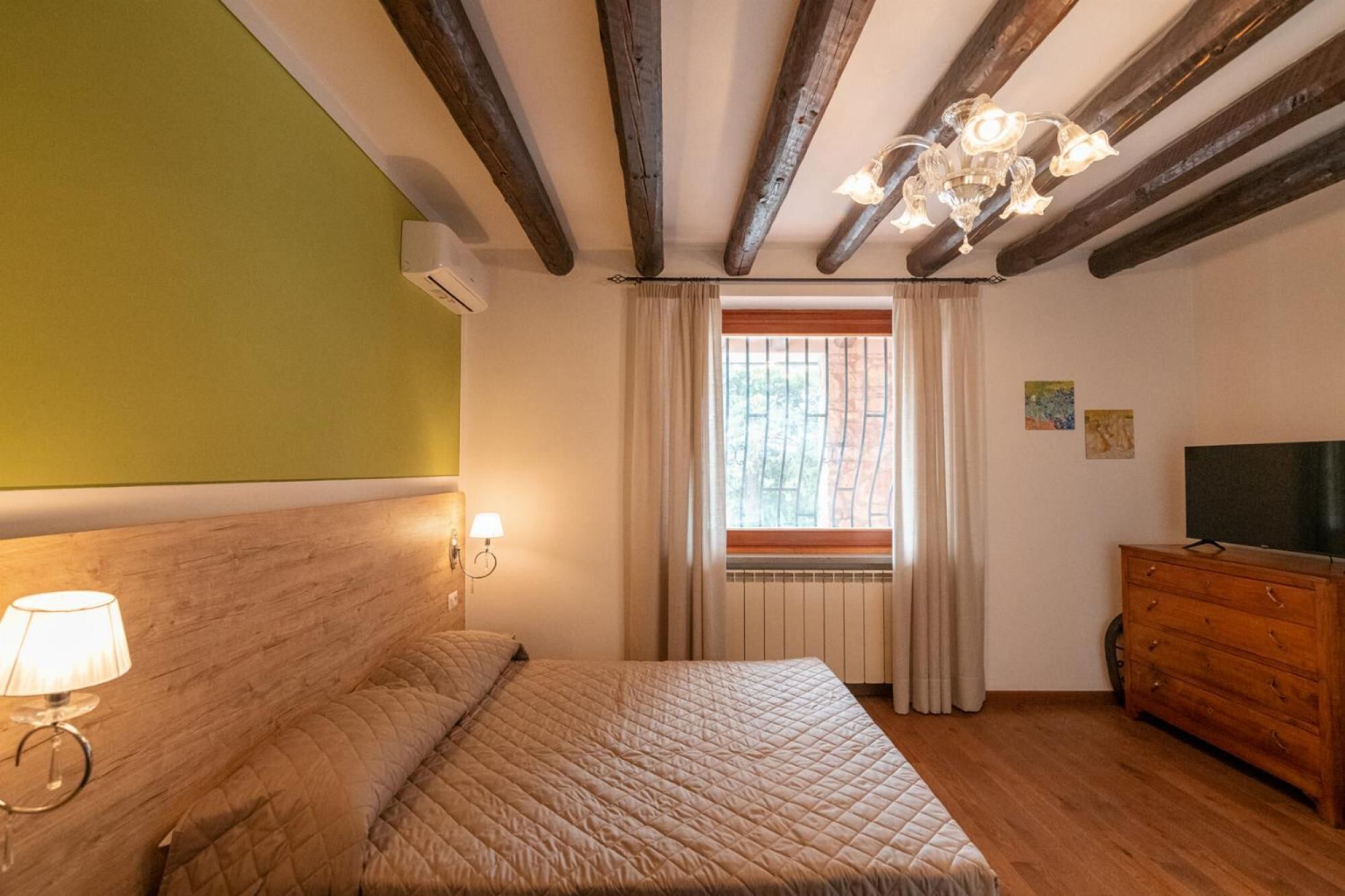 A bedroom with a bed, a nightstand with a lamp, a dresser, a window with curtains, and a chandelier.
