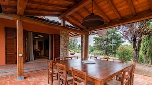 Um pátio externo com mesa e cadeiras de madeira, pilares de pedra e vista para o jardim ao redor.