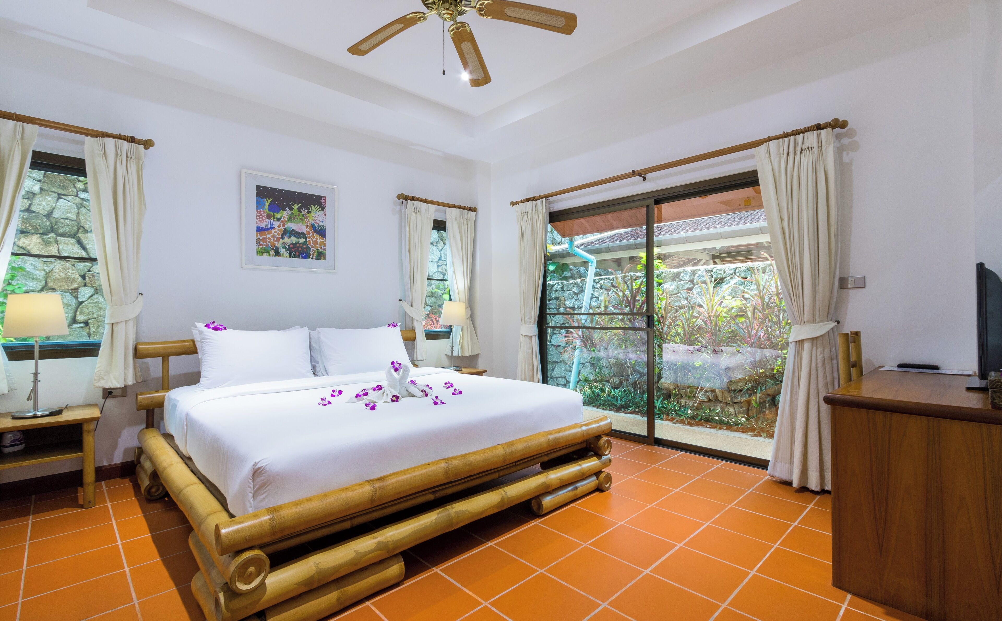A hotel room with a bed, a ceiling fan, a window with curtains, a wall-mounted TV, and a view of the outdoors.