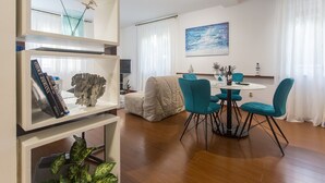 A modern living room with a round dining table, teal chairs, a white sofa, and a bookshelf with books and decorative items. - Top Location Beach & Downtown Split Apartment Park (Split)