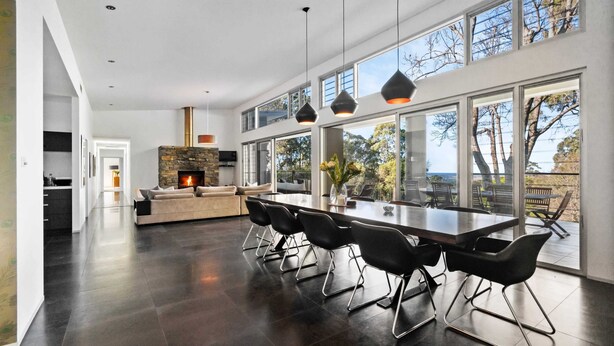 A modern kitchen with a stone fireplace, a dining area with black chairs, and large windows offering a view of the outdoors.