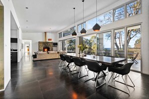 A modern kitchen with a stone fireplace, a dining area with black chairs, and large windows offering a view of the outdoors.