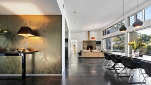 A modern kitchen with a stone fireplace, a dining area with a long table and chairs, and large windows offering a view of the outdoors.