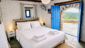 Ein Schlafzimmer mit einem Bett, weißen Bettwäsche, einem Nachttisch, einem Wandventilator, einem Fenster mit Blick auf Grünflächen und einem Steingewölbe.
