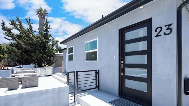 Un bâtiment moderne à la façade blanche, portant le numéro 23, avec une porte noire, des fenêtres et un petit espace extérieur aménagé avec des meubles en osier.