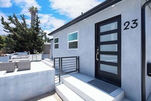Un bâtiment moderne à la façade blanche, portant le numéro 23, avec une porte noire, des fenêtres et un petit espace extérieur aménagé avec des meubles en osier.