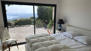 Un dormitorio con un ventanal grande, una cama, una silla y una mesita de noche con lámpara.