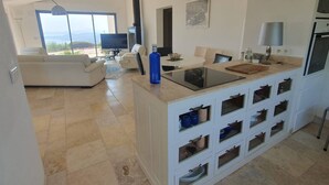 Una cocina moderna con una isla central, un comedor con televisor y una sala de estar con sofá y vistas al exterior.