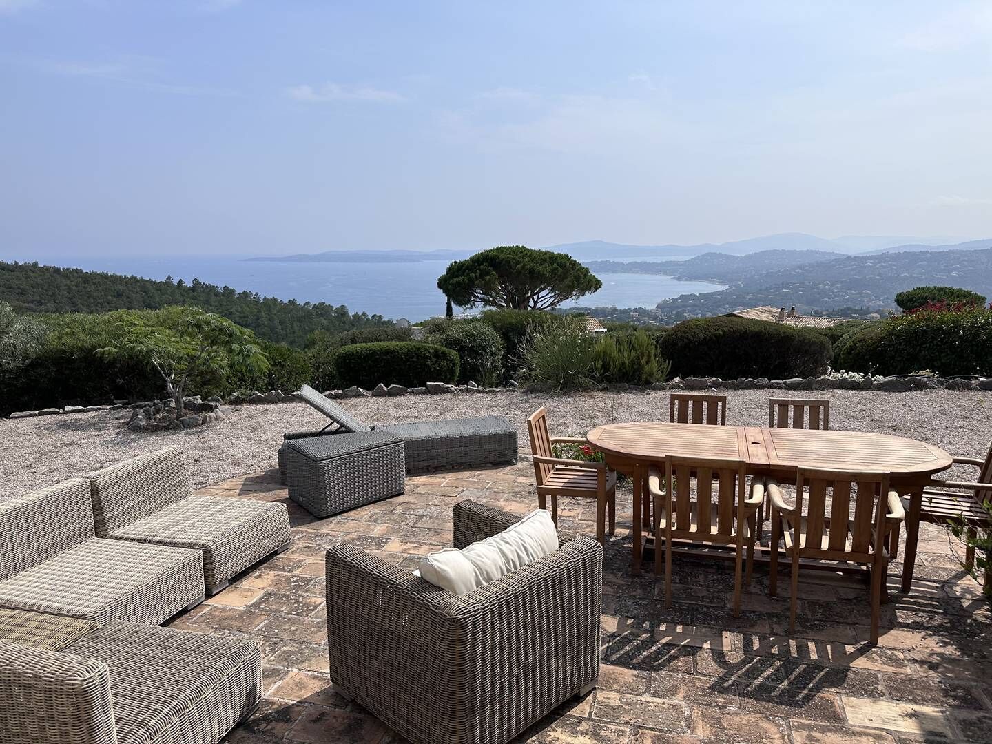 An outdoor seating area with wicker furniture and a wooden table, overlooking a scenic landscape with trees and a distant body of water.