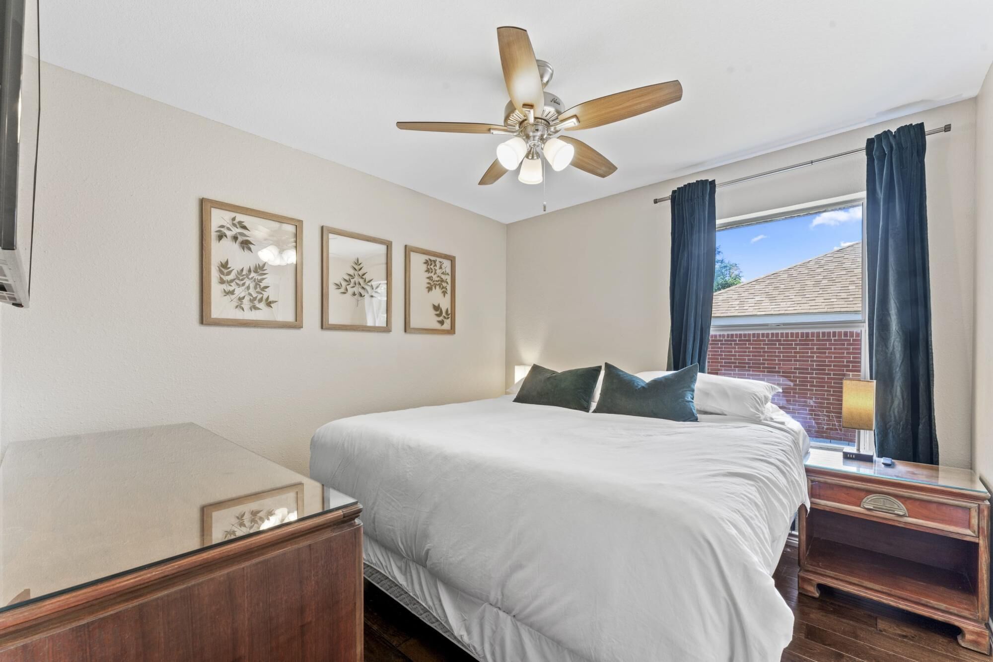 A bedroom with a bed, a ceiling fan, framed wall art, a nightstand, and a window with curtains.