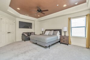 Une chambre à coucher comprenant un lit, des tables de chevet, un ventilateur de plafond, une fenêtre avec des rideaux et une porte.