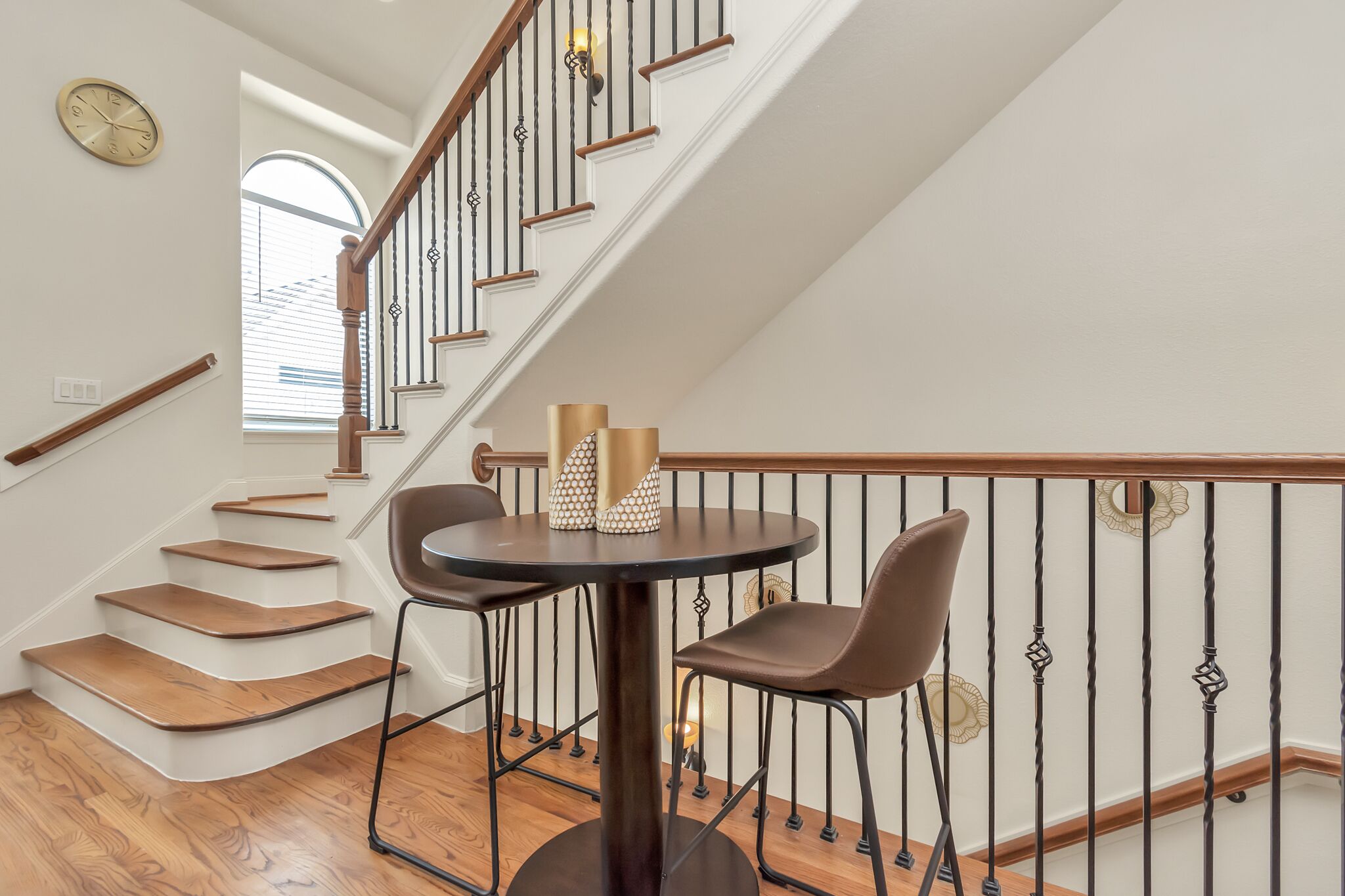Un escalier avec des marches en bois et une rampe noire, menant à un palier où se trouvent une table ronde et deux chaises.