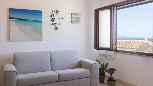 A living room with a beige sofa, a window with a view of the sea, and two potted plants. - Casa Su Lizu in Tanaunella (Budoni)