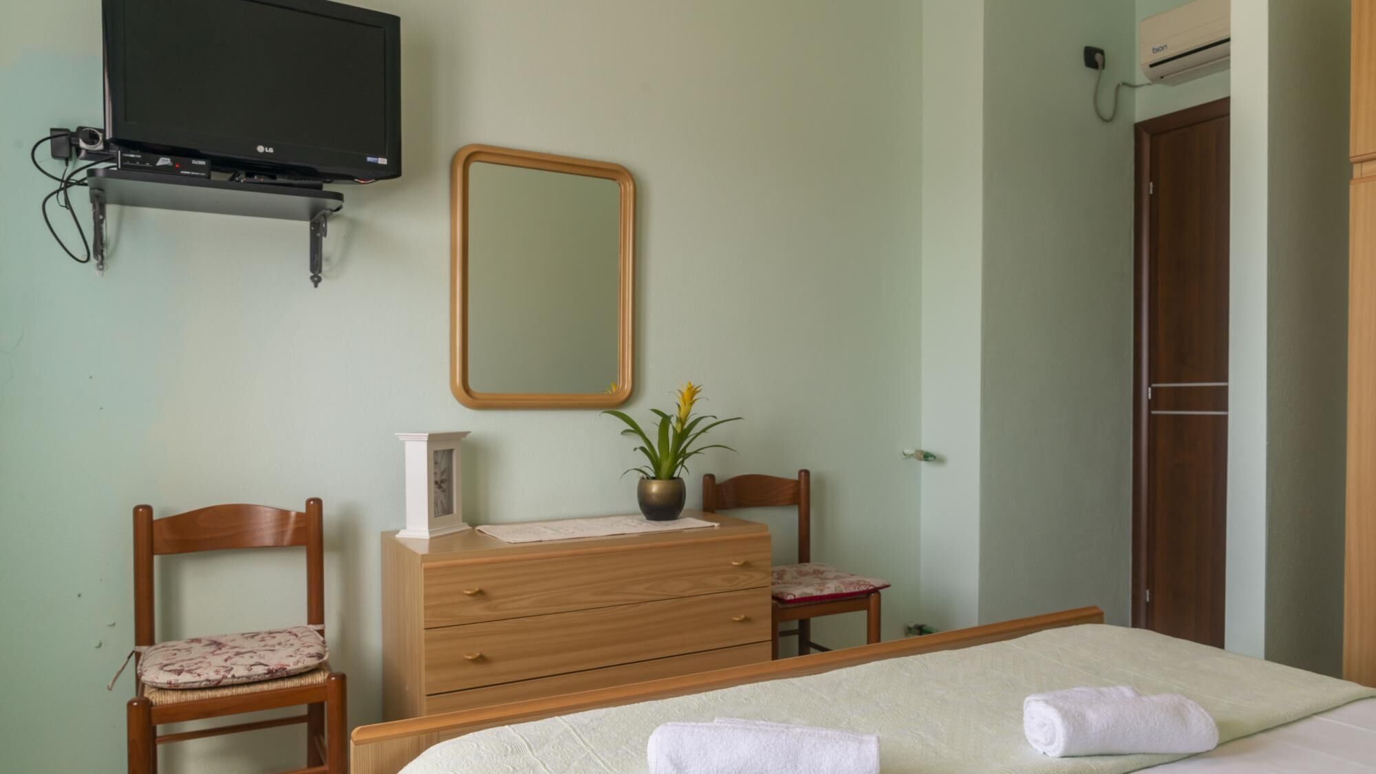 Une chambre à coucher comprenant un lit, deux chaises, une commode, un miroir et un téléviseur fixé au mur.