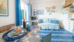 A room with a dining table set with food, a sofa with cushions, a bookshelf, and a window with curtains. - Gli Oleandri di Procchio (Procchio)