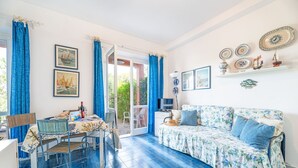 A room with a dining table, chairs, a sofa, and a TV. There are curtains, a window, and wall decorations. - Gli Oleandri di Procchio (Procchio)