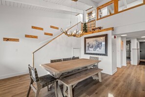 A spacious dining area with a wooden table and benches, a chandelier, and a staircase leading to an upper level.
