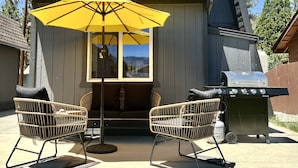 Een huis met een terras, waar twee rieten stoelen, een gele parasol en een barbecue staan.