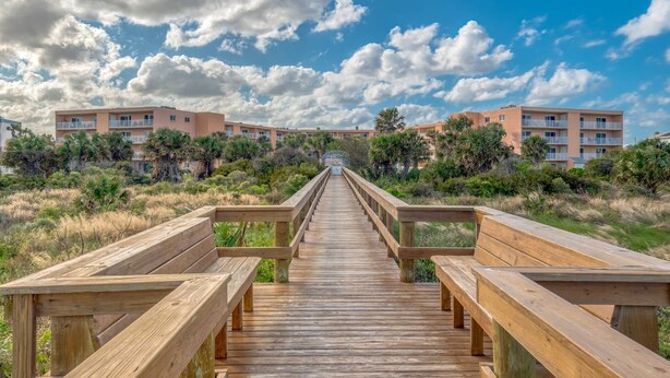 A wooden boardwalk leading to a building complex with balconies, surrounded by vegetation. - Amazing Ocean and Pool Views! Balcony, Elevator, Minutes to Historic Downtown, $300 in Free Tickets! (St. Augustine)