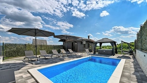 A rectangular swimming pool with a tiled edge, surrounded by lounge chairs and umbrellas, set against a backdrop of a clear sky and a hedge. - AquaHaven With Private Pool And Wellness (Veliko Trgovisce)