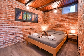 Un dormitorio con pared de ladrillo, una cama con colcha gris, dos mesitas de noche con lámparas y un cuadro abstracto enmarcado.
