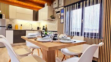Une cuisine moderne avec des meubles blancs, une table à manger en bois pour quatre personnes et une vue par une fenêtre équipée de stores verticaux.