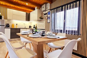 A modern kitchen with white cabinets, a wooden dining table set for four, and a view through a window with vertical blinds. - AquaHaven With Private Pool And Wellness (Veliko Trgovisce)