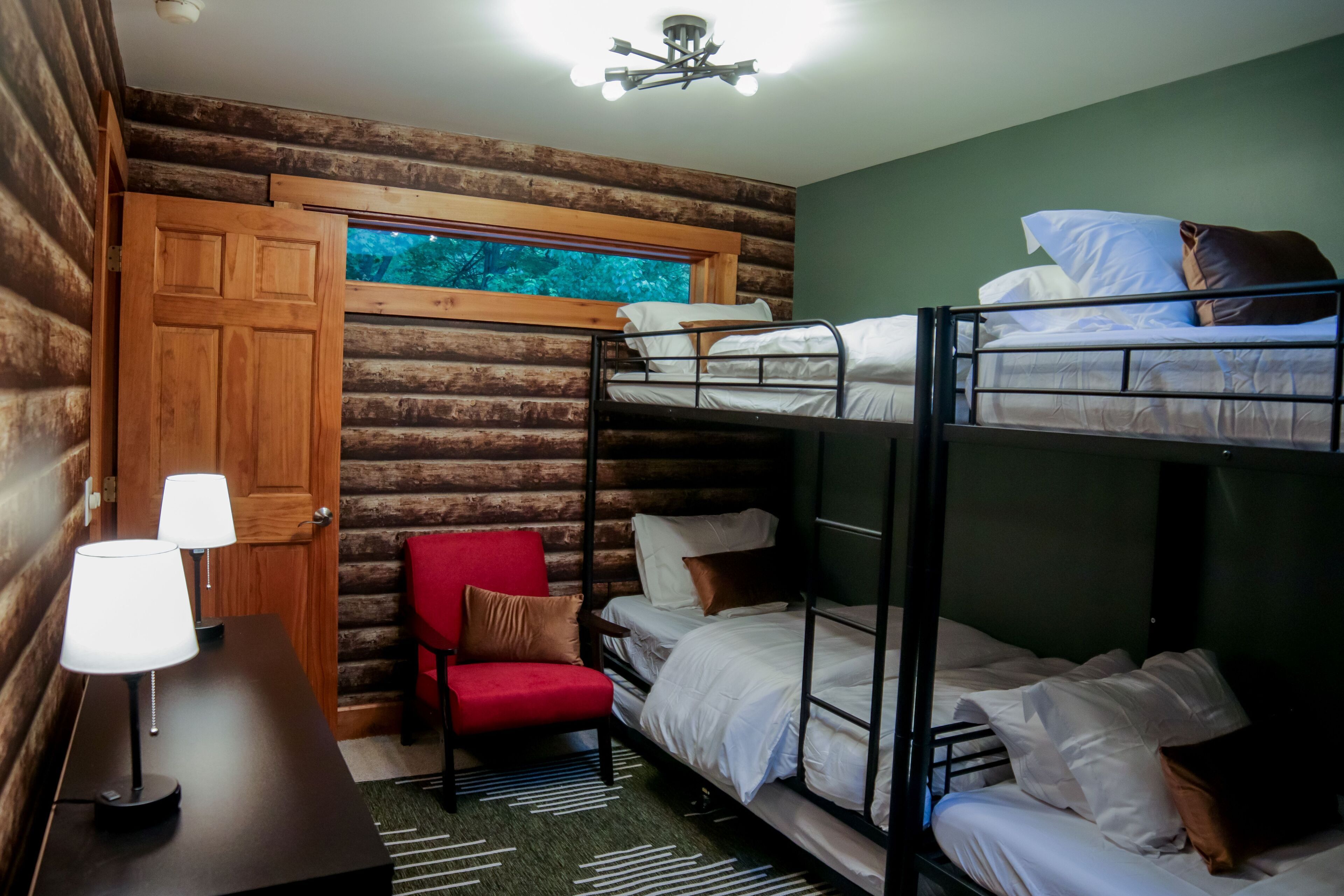 A bunk bed room with a wooden door, a red chair, a table with a lamp, and a window with a view of trees.
