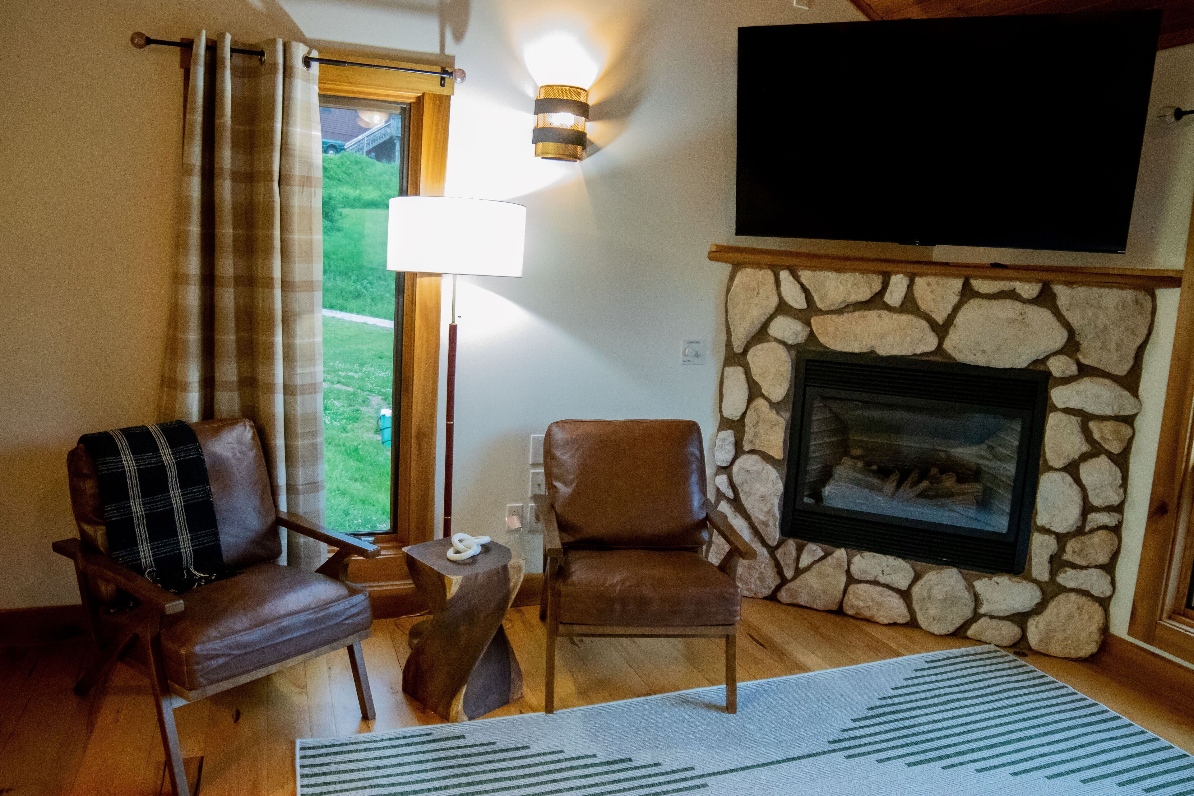 Un salon chaleureux avec une cheminée en pierre, des fauteuils en cuir, un téléviseur à écran plat et une grande fenêtre agrémentée de rideaux.