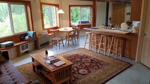 A cozy living room with a sofa, a coffee table, and a rug. There is a dining area with a table and chairs, and a kitchen area with a counter and stools. - Charming house with orchard and pond close to Flathead Lake (Rollins)