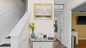 A hallway with a white staircase, a welcome sign, a framed mirror, and a door leading to another room. - Beautiful Luxury Budget Home With Pool. Near 6Flags / AT&T (Grand Prairie)