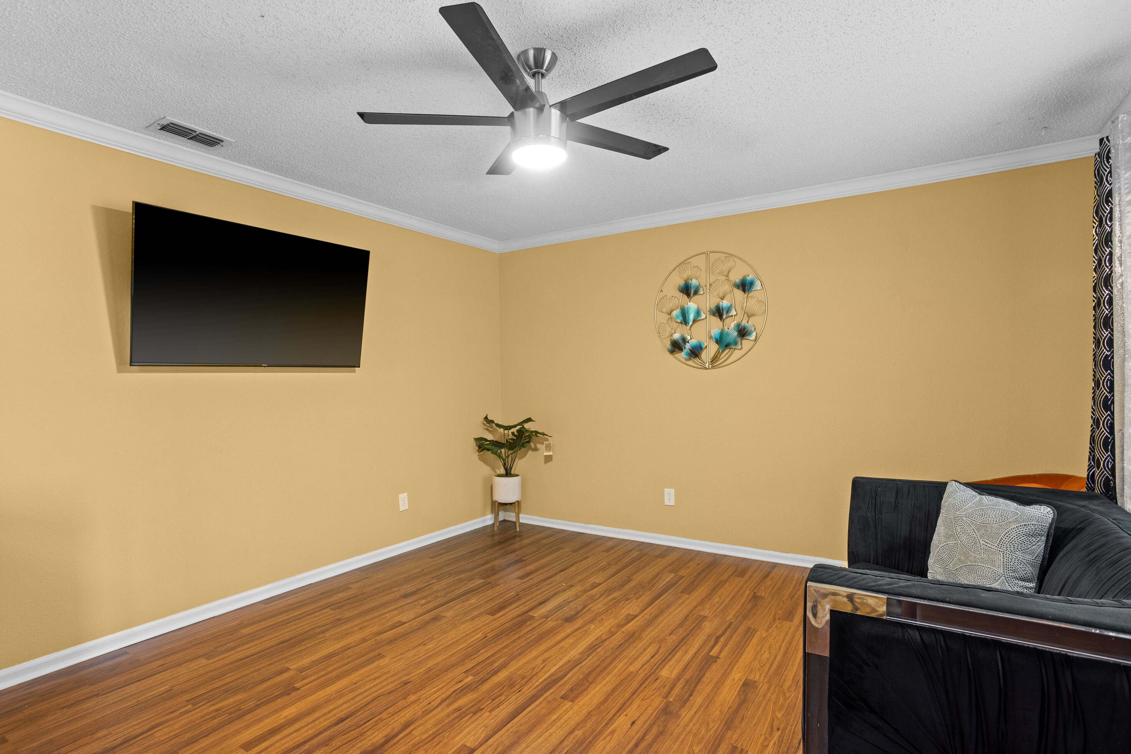 Quarto com ventilador no teto, televisão fixada na parede, um objeto decorativo, uma planta em vaso e um sofá preto com almofada.