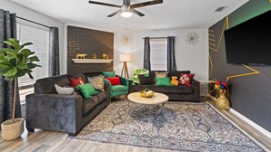 A modern living room with two sofas, a coffee table, a ceiling fan, a fireplace, and a large TV. - Beautiful Luxury Budget Home With Pool. Near 6Flags / AT&T (Grand Prairie)