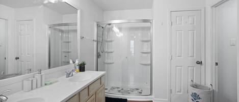 A modern bathroom with a large mirror, two sinks, a shower enclosure, and a door.