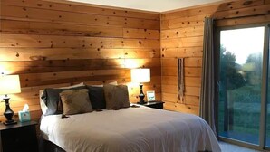 A bedroom with a wooden paneled ceiling, a large bed with white bedding, two bedside tables with lamps, and a sliding glass door leading to a balcony.