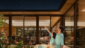 Un área de piscina interior moderna con barandillas de vidrio, rodeada de plantas exuberantes y un cielo nocturno estrellado visible a través de amplias ventanas.