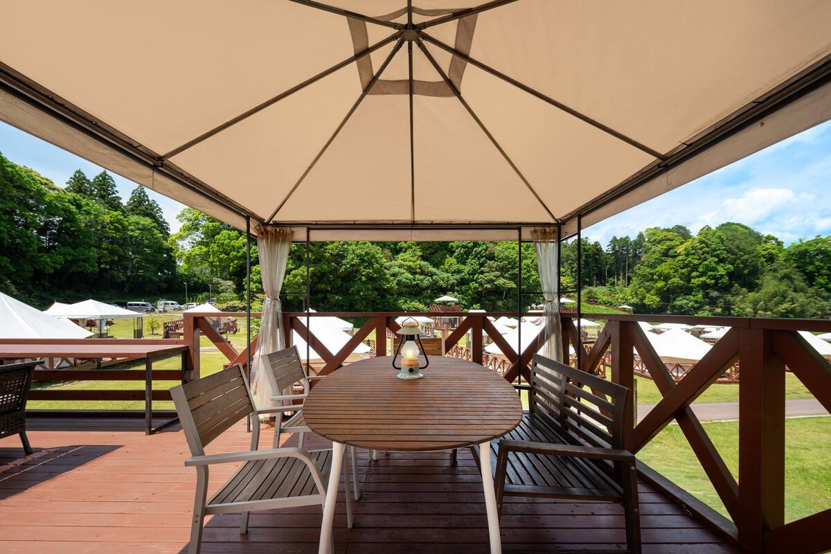 Un espacio de estar al aire libre cubierto con una mesa y sillas, rodeado por una terraza de madera y vistas a un paisaje verde exuberante.