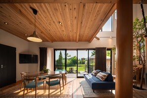 Uma sala de estar moderna com teto de madeira, sofá, mesa de jantar com cadeiras e uma grande janela com vista para o exterior.