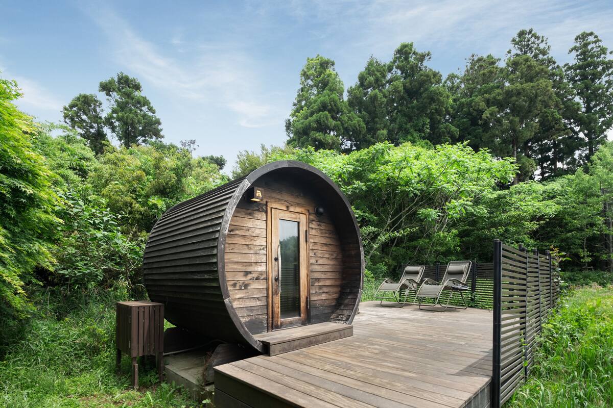 Una cabaña de madera cilíndrica con una puerta, un taburete de madera y dos sillones de descanso en una terraza de madera rodeada de vegetación.