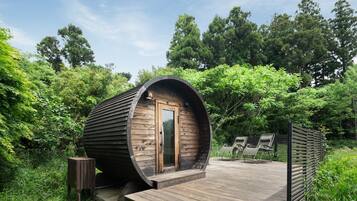 Een houten vakantiehuisje in cilinder vorm, met een deur, een klein houten krukje en twee comfortabele stoelen op een houten terras, omringd door groen.