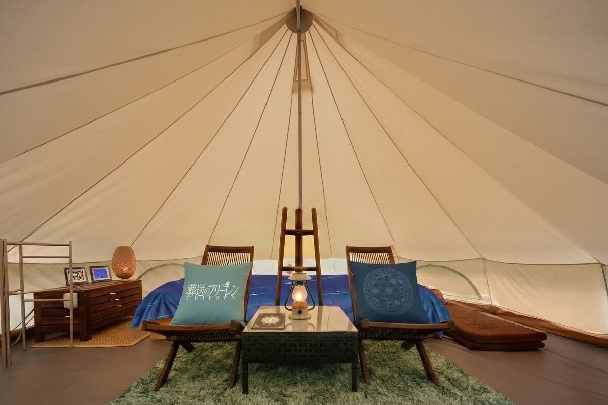 Una carpa con una mesa central, dos sillas, una escalera y un pequeño escritorio con una lámpara.