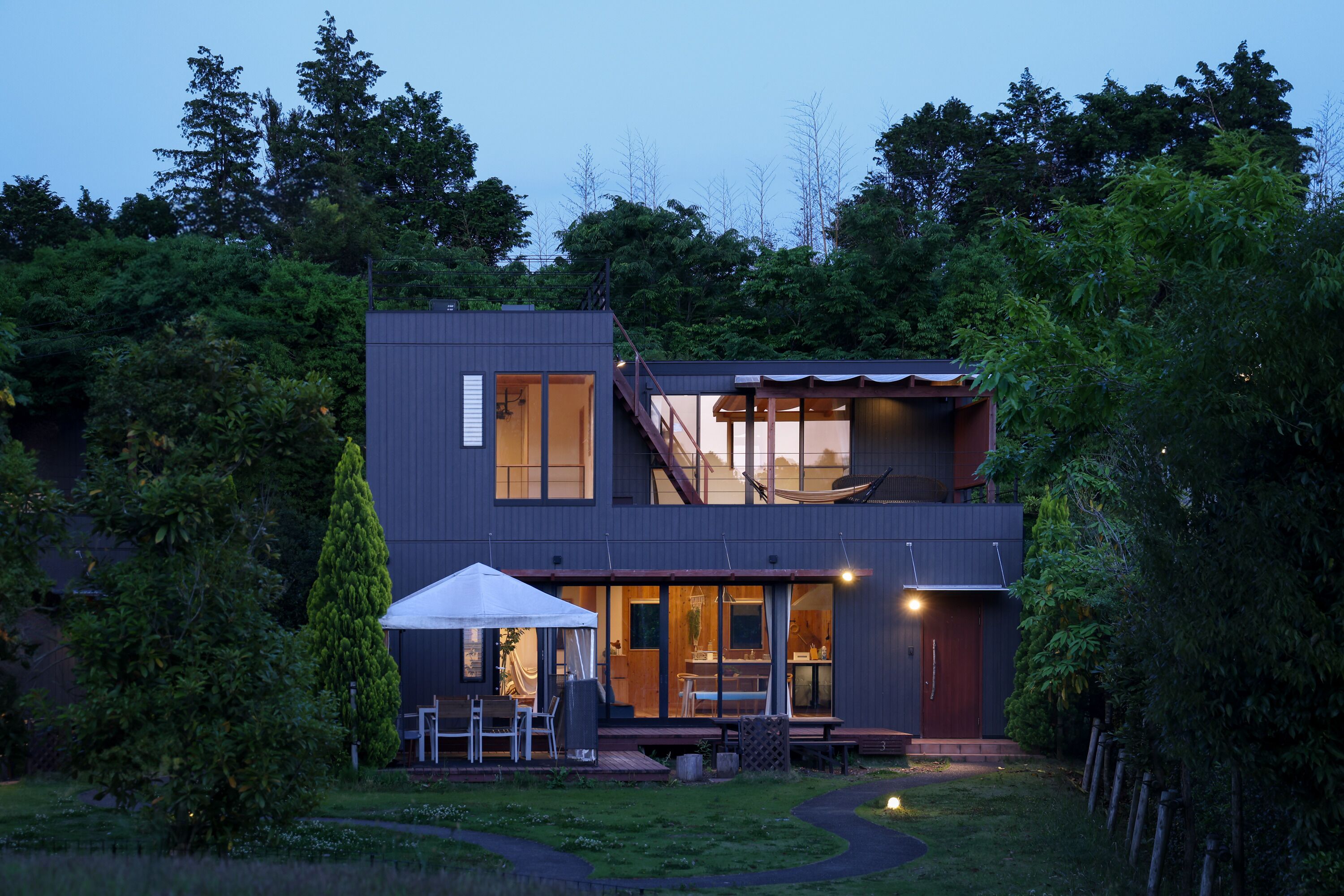 Ein modernes, zweistöckiges Haus mit überdachter Terrasse, großen Fenstern und einer Treppe, die zu einer oberen Ebene führt.