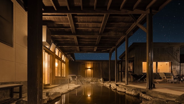 Zona de aguas termales nocturna con techo de madera, asientos de piedra y vista al cielo estrellado.